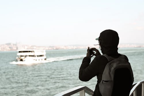 Back view of unrecognizable man with backpack in jacket admiring ship in wavy sea using small camera