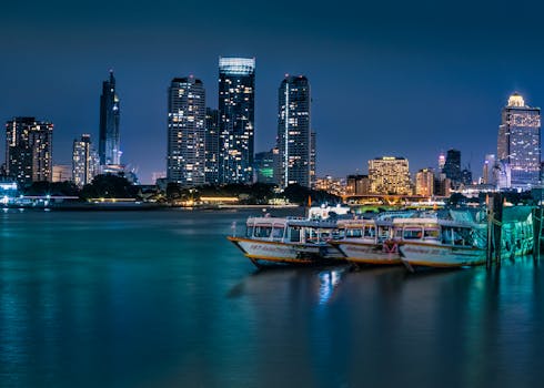 Illuminated city skyline with skyscrapers and boats on a waterfront at night.