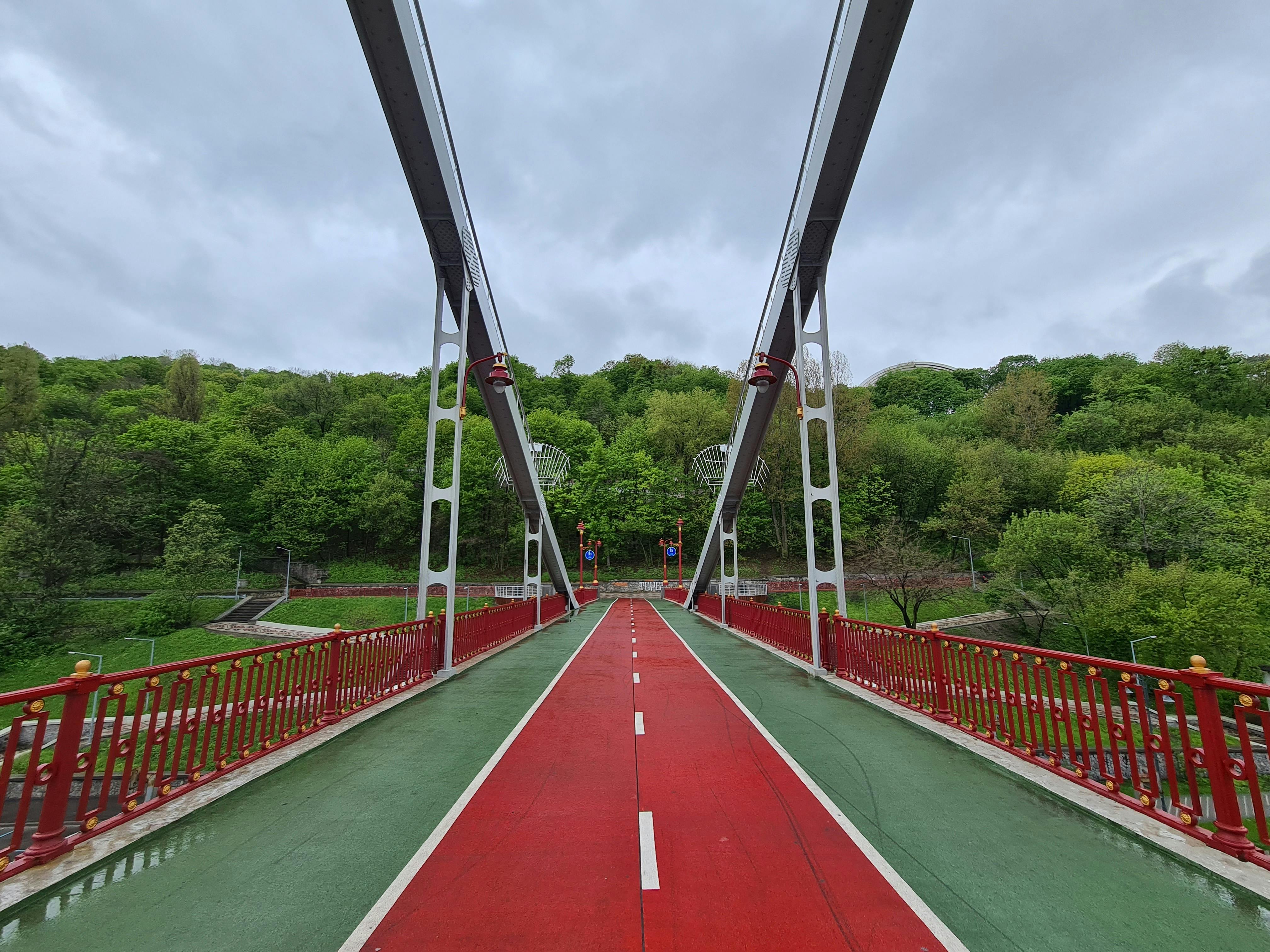 Sight of a red pedestrian bridge amidst lush green trees under a cloudy sky.