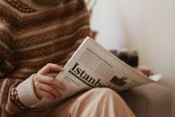 Crop unrecognizable female in ornamental sweater reading newspaper article while sitting in armchair in house room