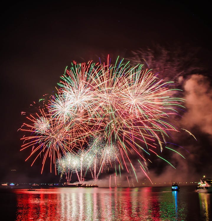 Free Colorful fireworks illuminating the night sky above a serene water body, creating a festive atmosphere. Stock Photo