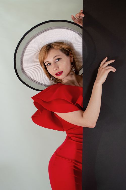 Free Stylish woman in a red dress with a wide-brimmed hat posing against a black panel. Stock Photo
