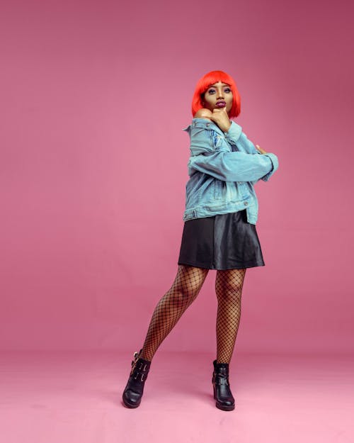Free Full body of young African American female with dyed hair wearing denim jacket and mesh tights looking at camera on pink background in studio Stock Photo