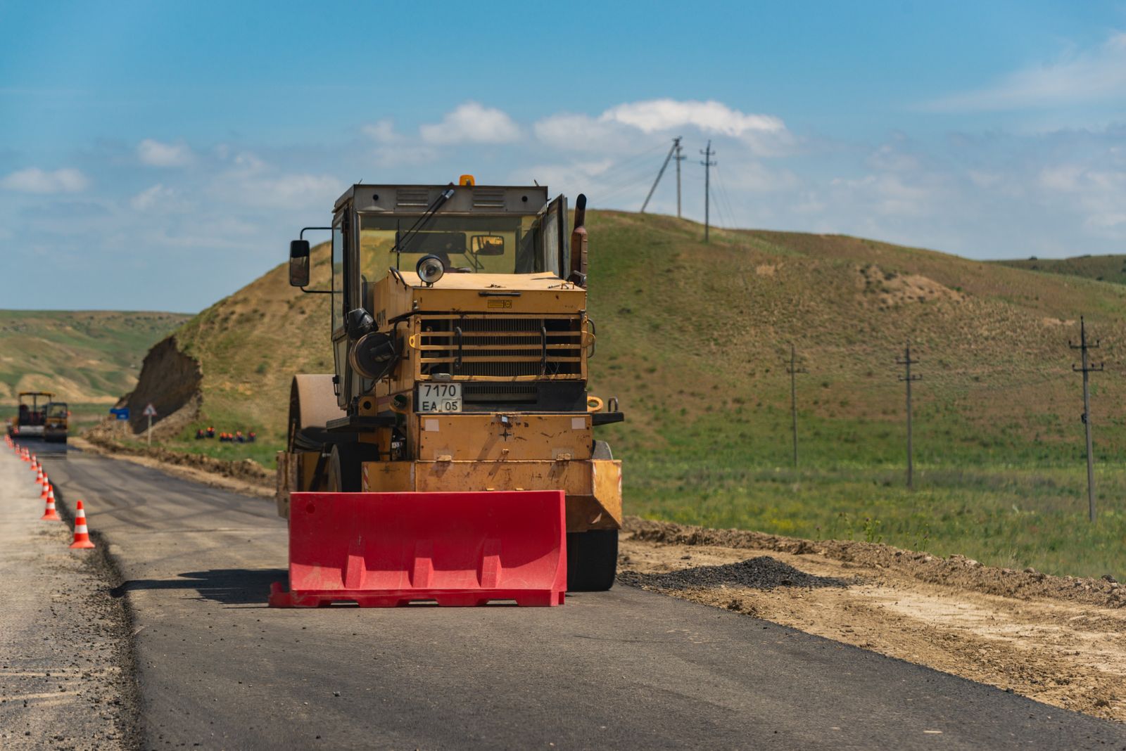 Un rodillo compactador amarillo sobre una pista recién asfaltada en el campo, con conos de seguridad a un lado, cerros verdes y postes de luz al fondo