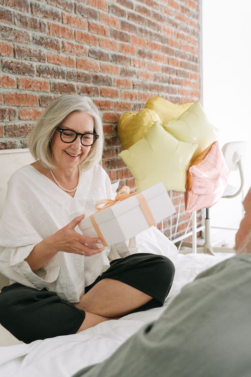 Free Joyful senior woman opening a gift with balloons in a cozy indoor setting, celebrating a special occasion. Stock Photo