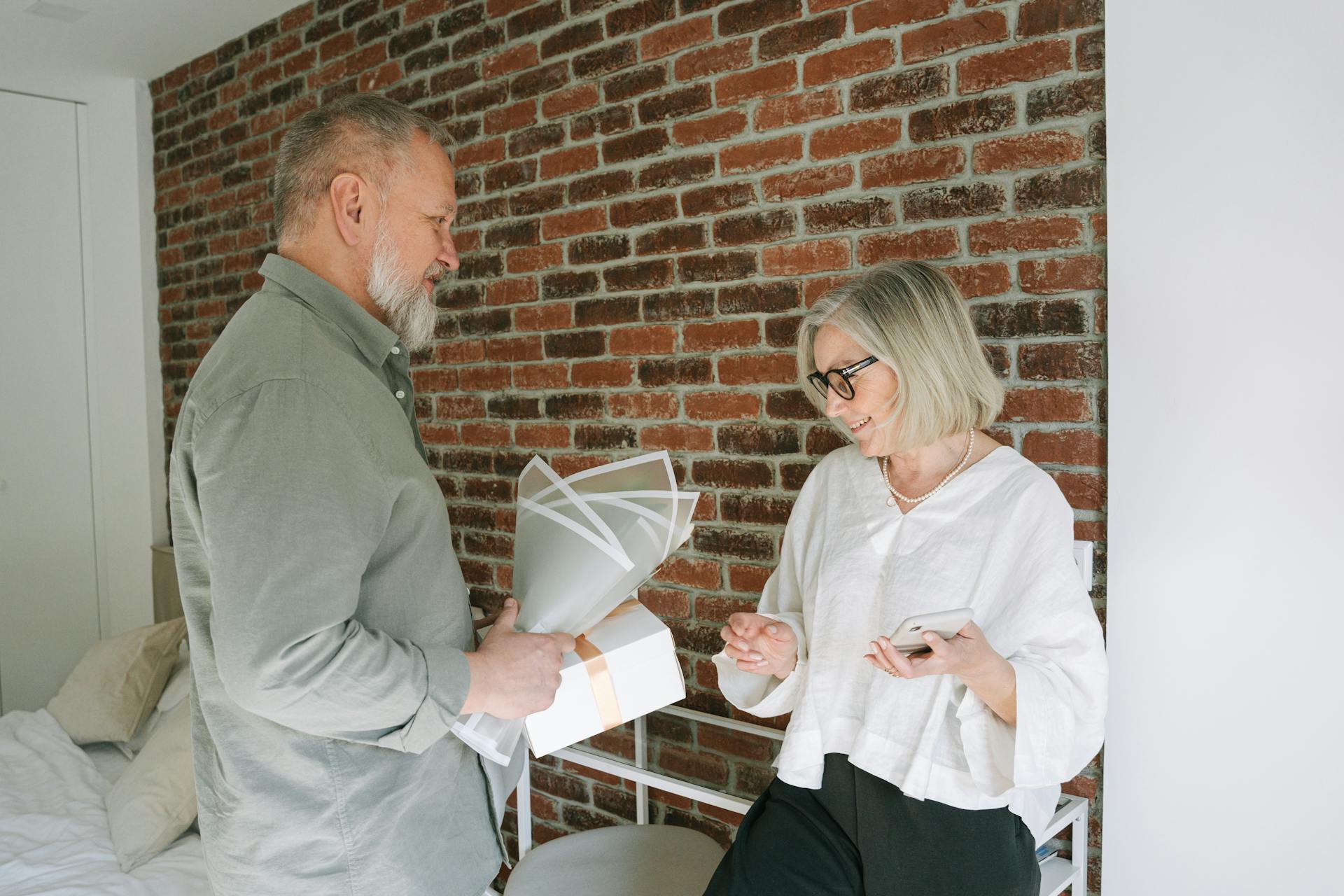 A senior couple shares a romantic moment with a gift against a brick wall background indoors.