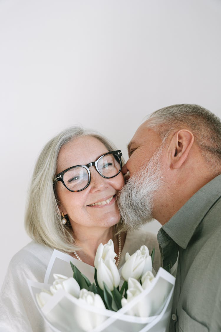 Senior Man In Gray Shirt Kissing His Wife On Her Cheek While Holding A Bouquet Of Flowers