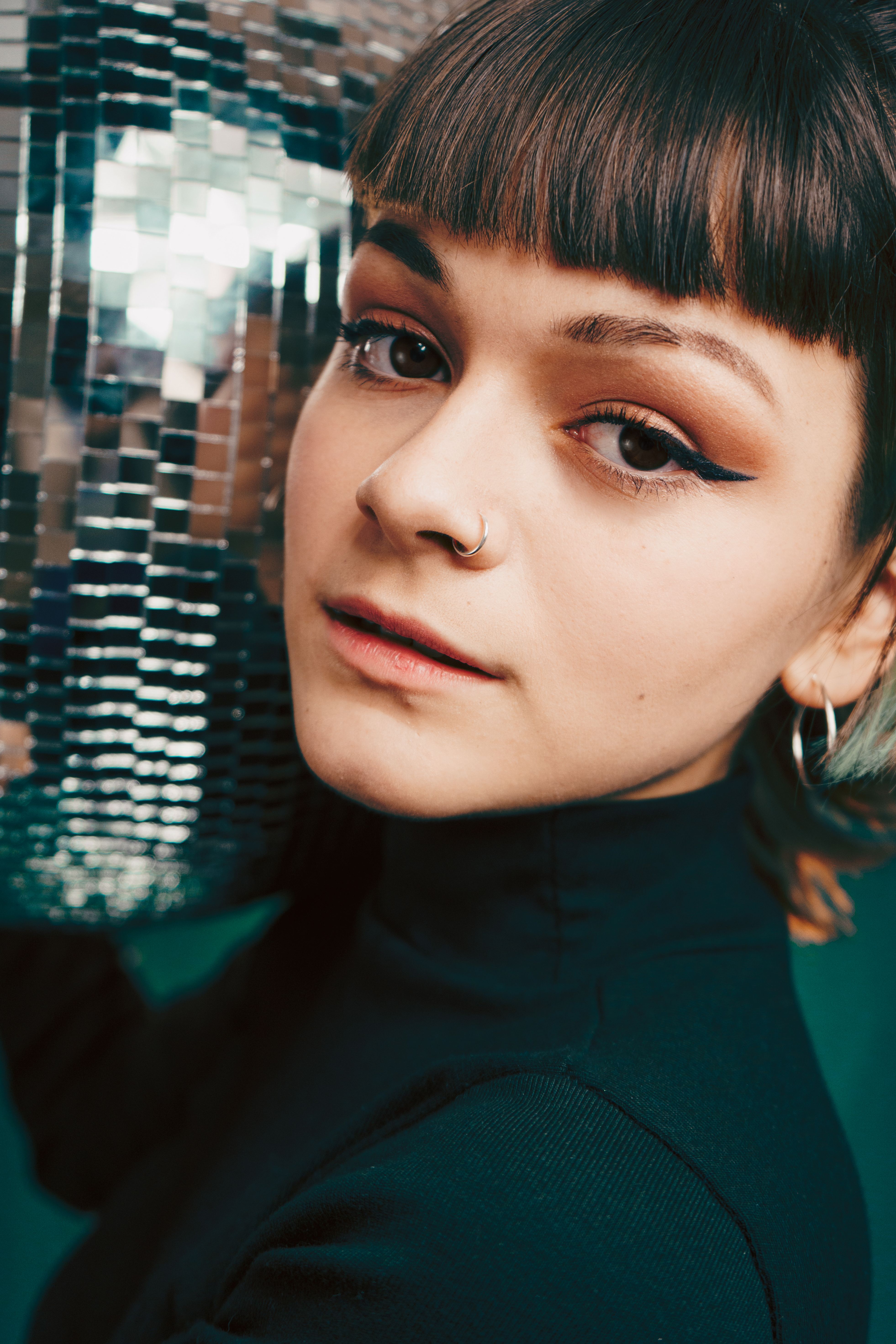 Close-up portrait of a woman with bold eyeliner and hoop earrings, leaning on a disco ball.