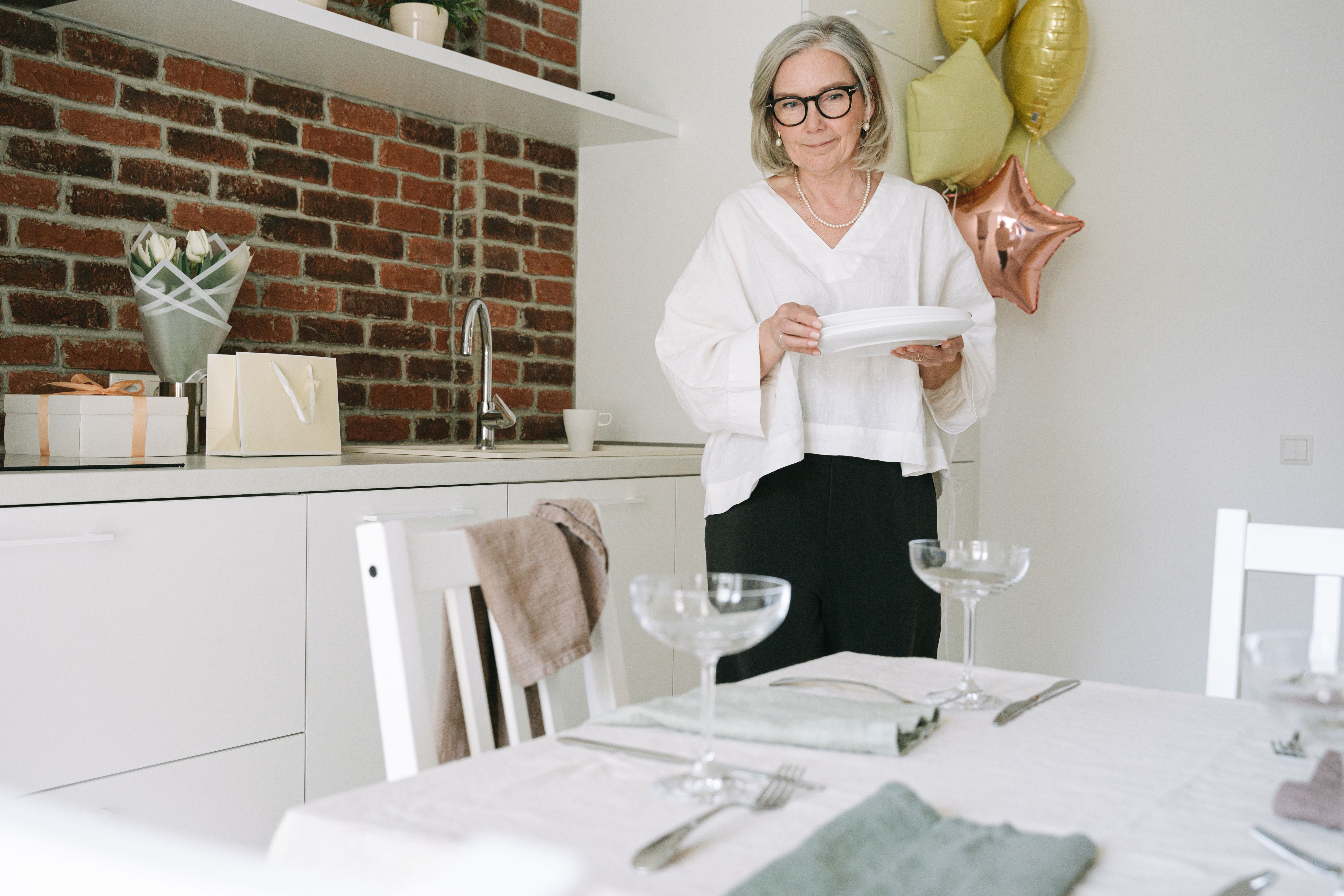 Elderly woman setting a dining table with plates and glasses in a cozy home kitchen.