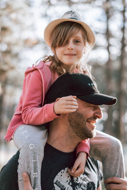 Delighted father in cap giving ride on shoulders to cute daughter while spending weekend together in park