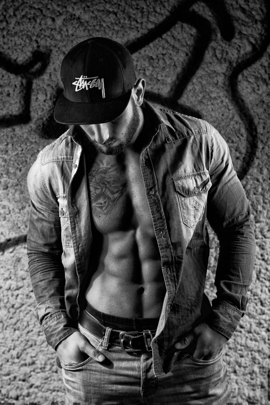 A muscular man wearing a denim shirt and cap poses against a graffiti-covered wall, showcasing his physique.