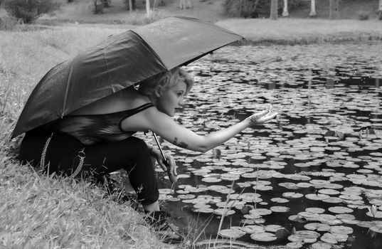 A woman sitting under an umbrella by a pond filled with water lilies during a rain shower.