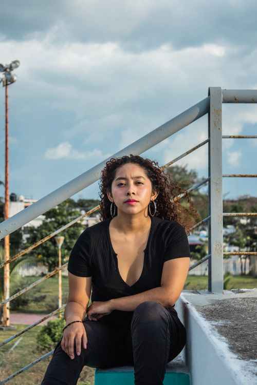 Free Woman with curly hair seated outdoors on steps, featuring a rusty handrail and cloudy sky. Stock Photo
