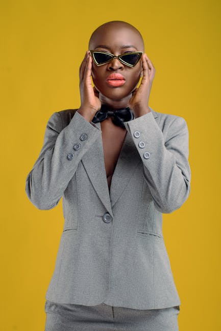 Trendy African American female with lipstick wearing elegant gray suit and bow tie standing on yellow background in light studio