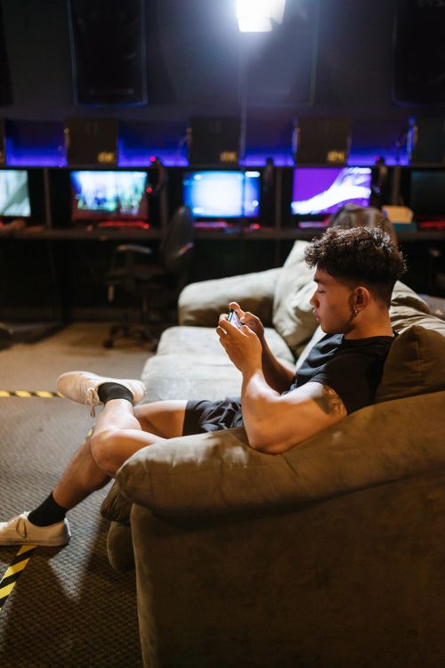 Free A young man in casual attire relaxes on a sofa playing games on his smartphone at an internet cafe. Stock Photo