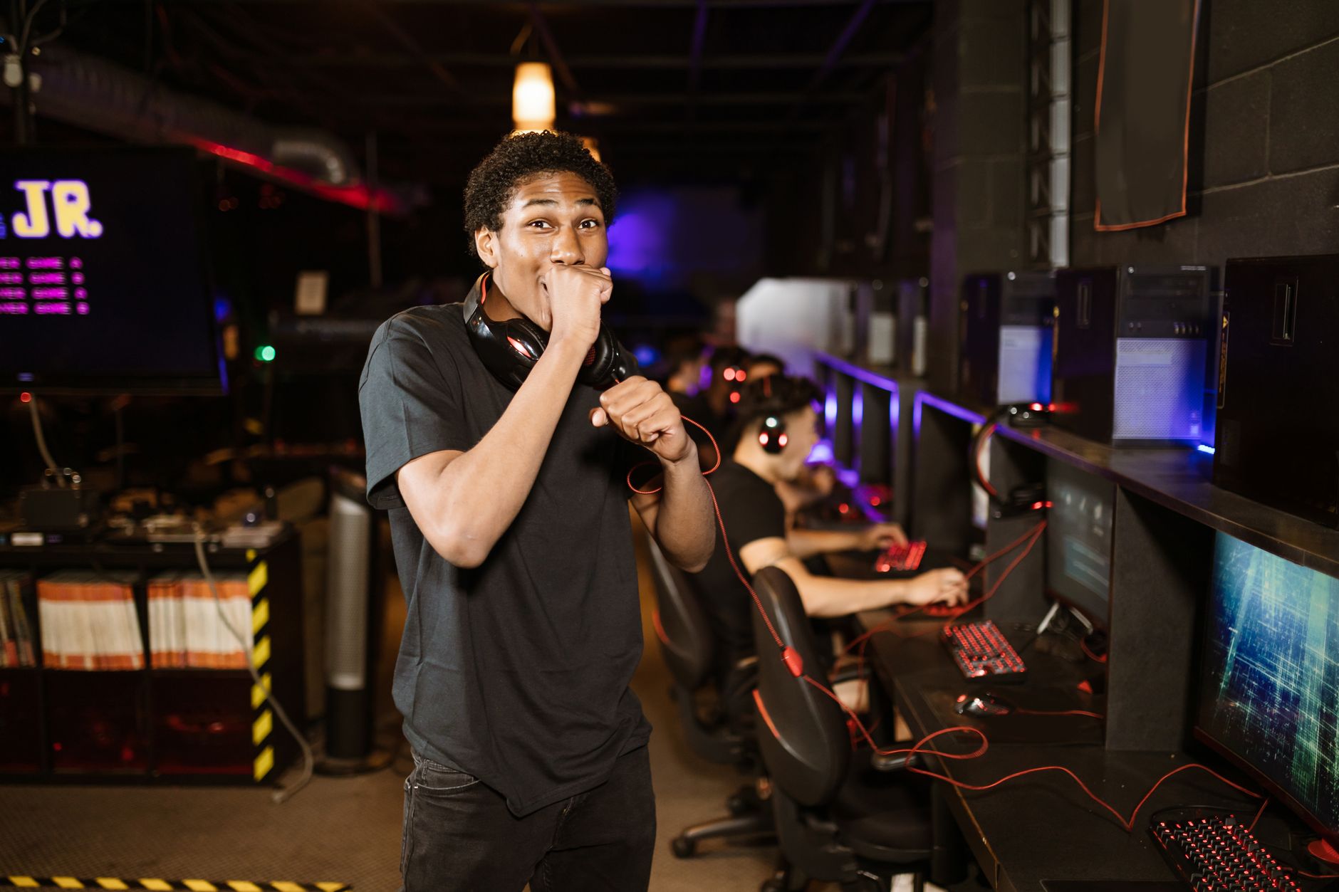 A young gamer excitedly posing in a dimly lit esports arena. Others playing in the background.