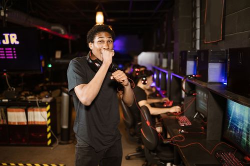 Free A young gamer excitedly posing in a dimly lit esports arena. Others playing in the background. Stock Photo