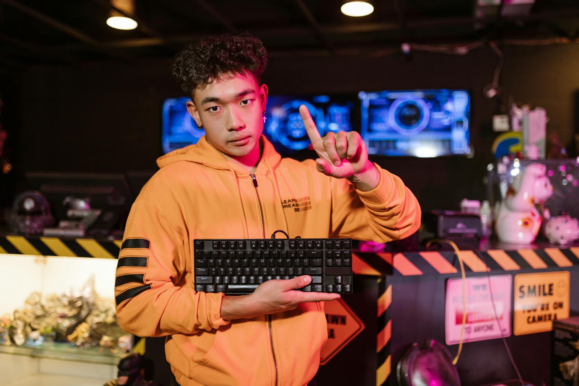 A young man in an orange jacket poses with a mechanical keyboard in a gaming setup.