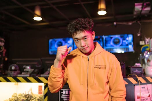 Young Asian male gamer standing confidently indoors wearing an orange jacket.