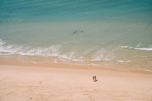 A serene aerial shot of a sandy beach with two people walking along the shore.