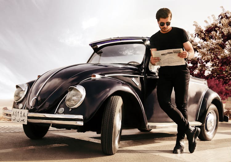 A Man Reading A Newspaper Standing Beside A Vintage Car