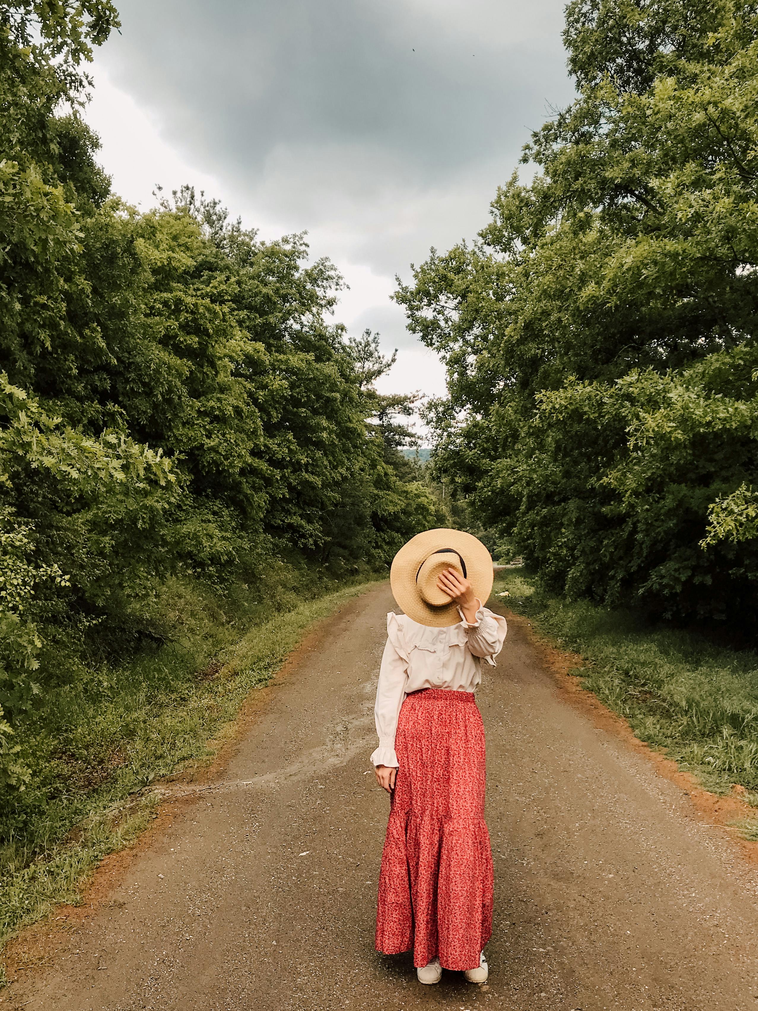 Full body of unrecognizable female in stylish outfit covering face with hat while standing on walkway in forest on cloudy day