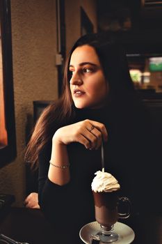 A woman sits indoors enjoying a chocolate shake with whipped cream, captured in warm lighting.