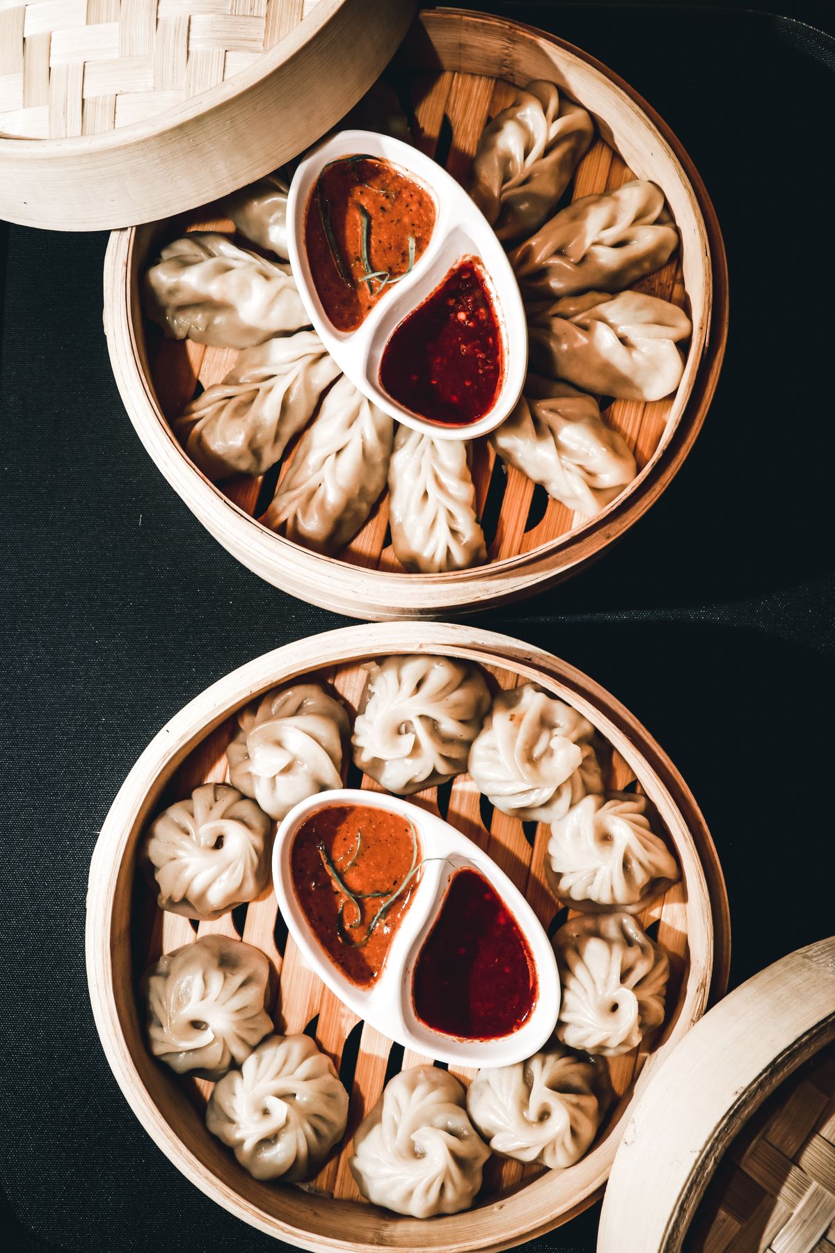 Steamed momos packed into two bamboo baskets in Nepal, dishes of red and orange chilli chutney between them