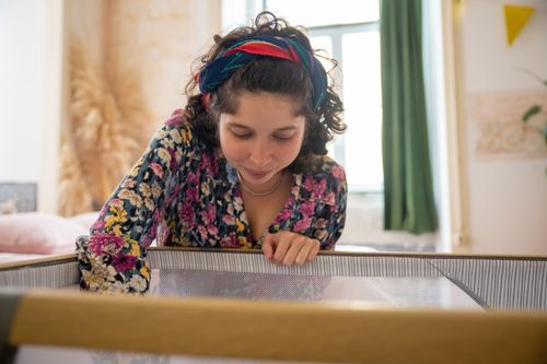 Free A joyful woman with a floral shirt gazes into a crib, creating a warm home atmosphere. Stock Photo