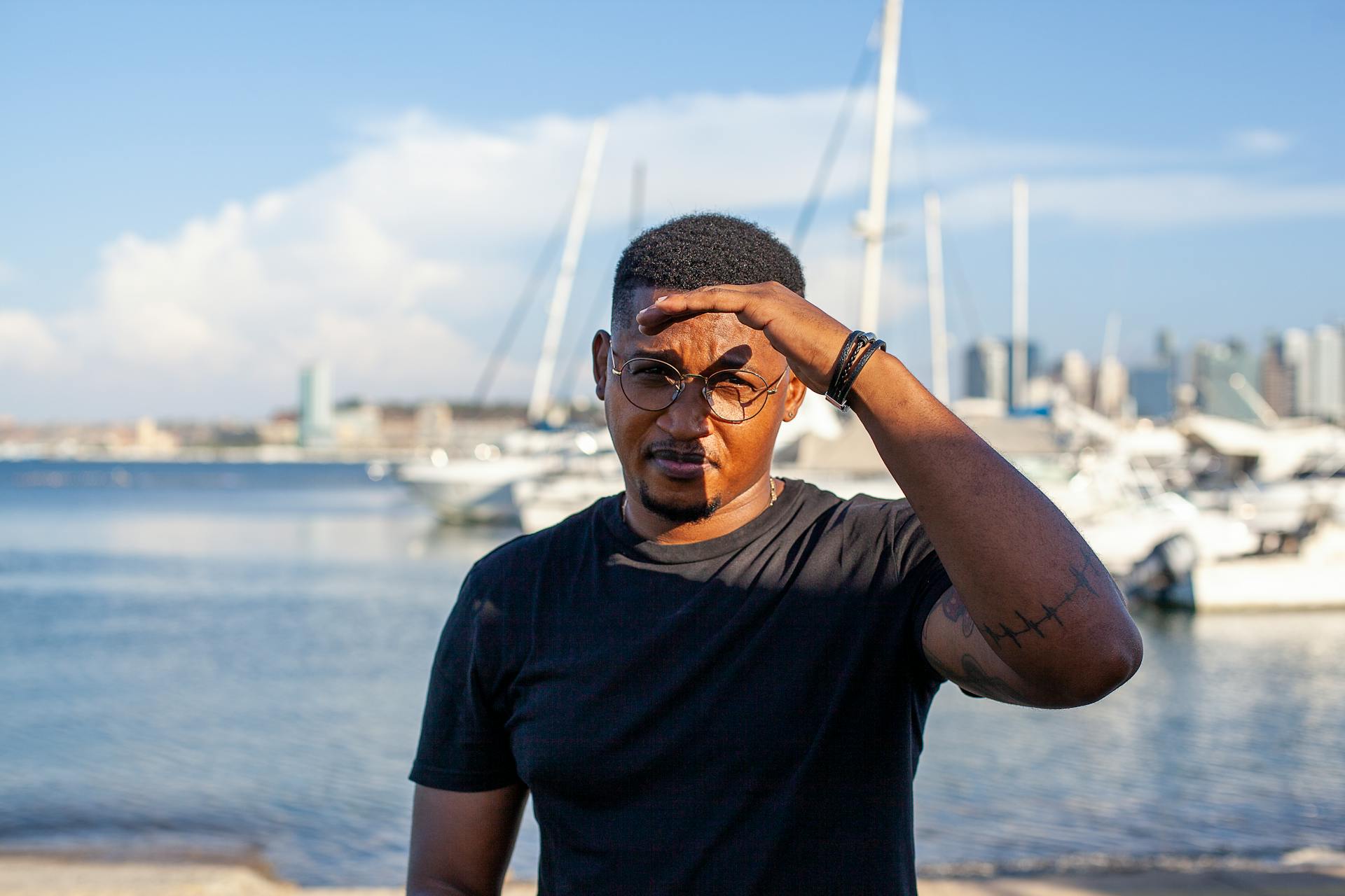 Man in glasses shielding eyes at Luanda harbor with yachts in the background.