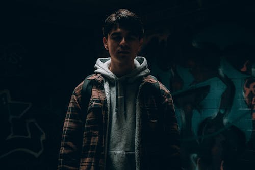 Free Young Man in Gray Hoodie with Plaid Jacket Standing Near a Wall with Graffiti Stock Photo