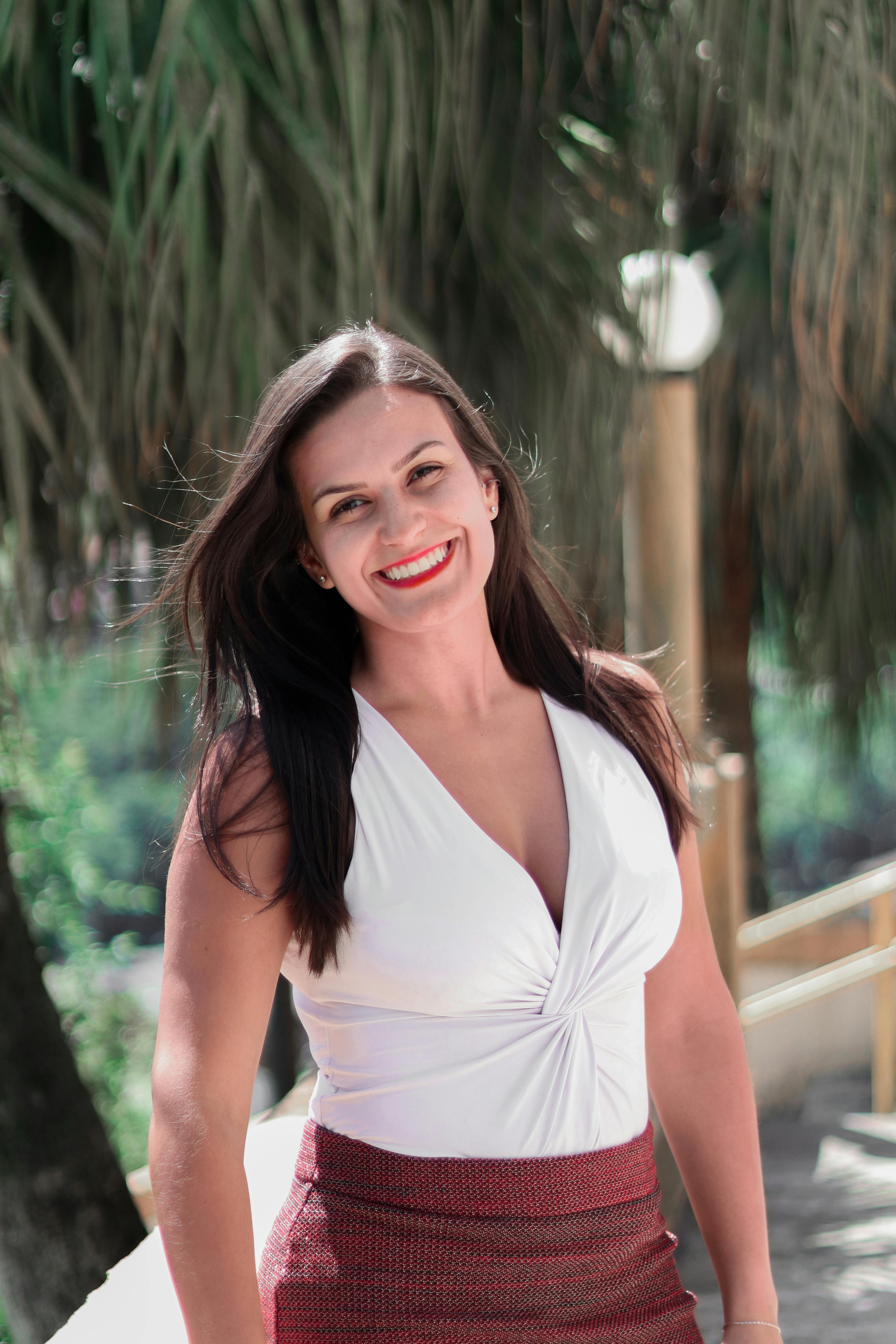 Happy woman in a white top smiling brightly outdoors with trees in background.