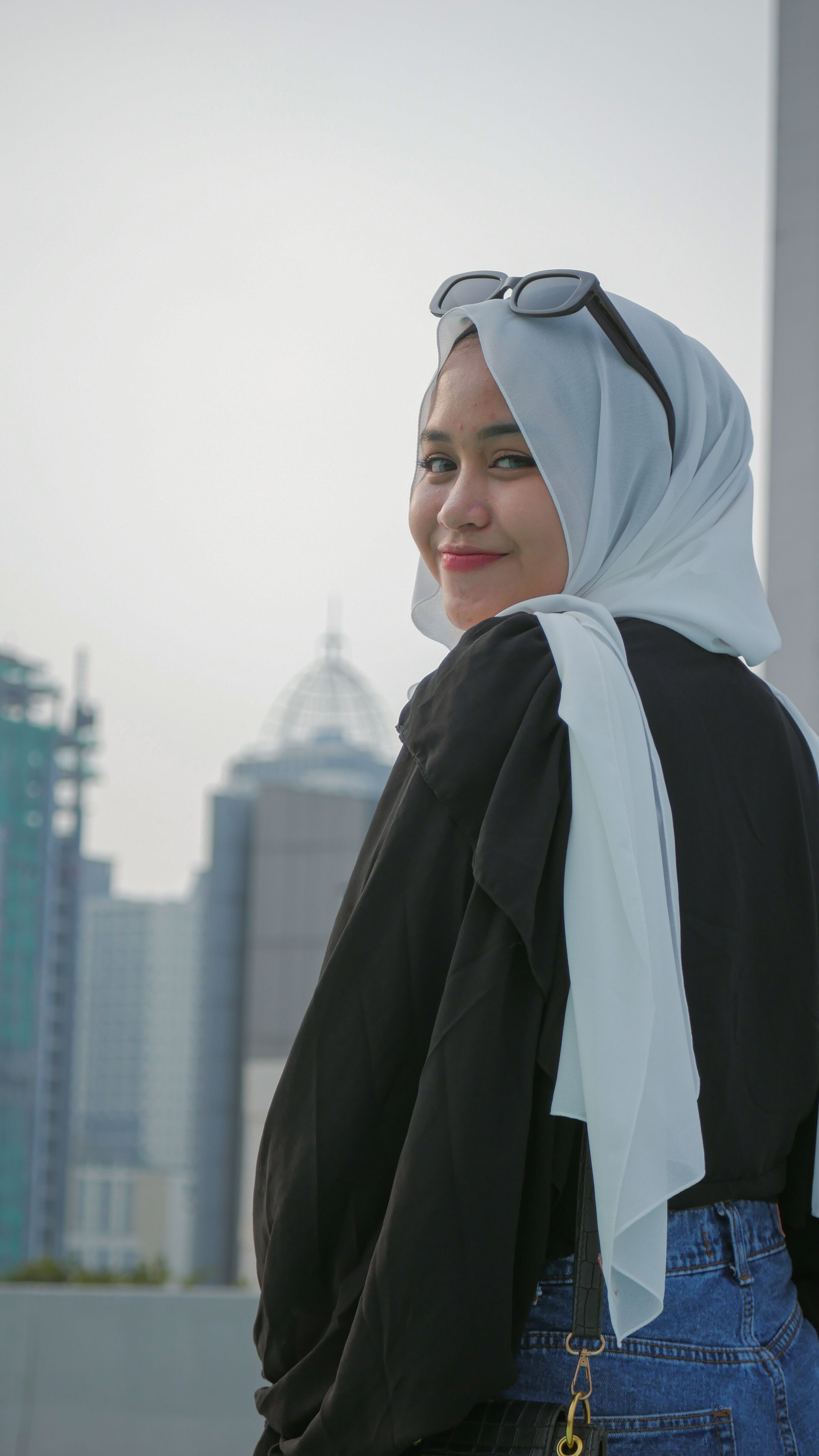 A smiling woman in a headscarf stands outdoors in a city, exuding confidence and style.