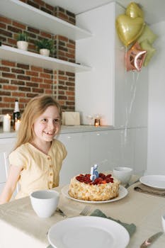 Smiling girl celebrating her birthday with a decorated cake and colorful balloons indoors.