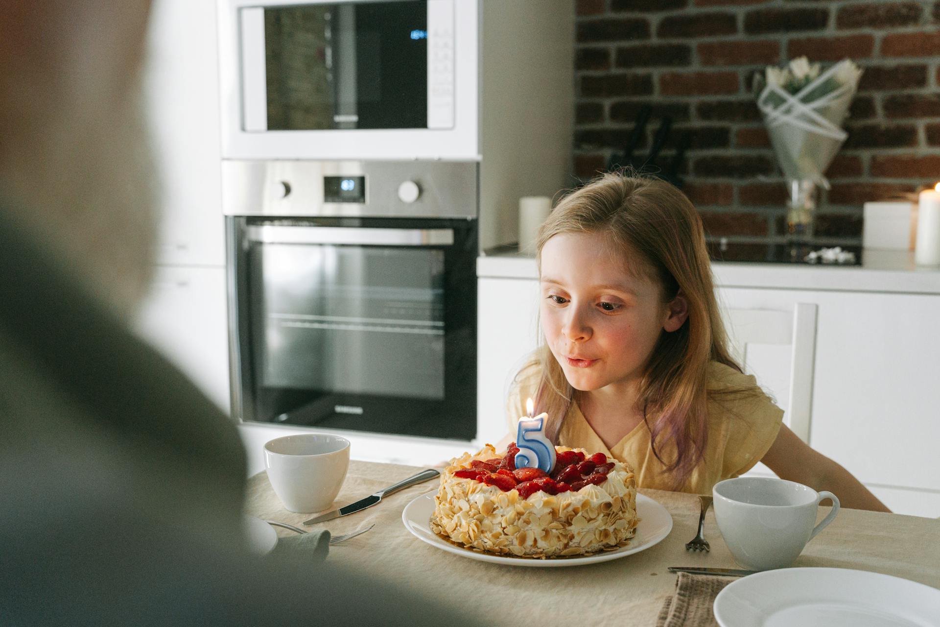 A young girl blows out a birthday candle on a cake during a fifth birthday celebration indoors.