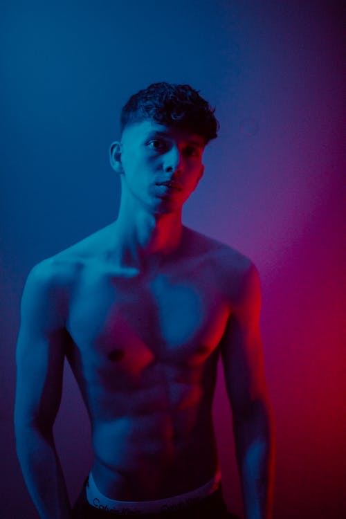 Muscular young male with naked torso looking at camera while standing near wall in dark room with glowing neon illumination