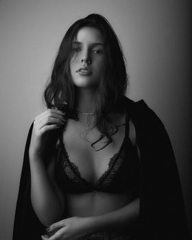 Black and white of young woman with dark hair in lace lingerie and jacket on shoulders looking at camera against light wall in room
