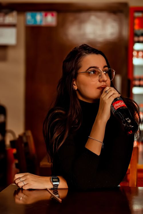 Free Woman wearing glasses sipping Coca-Cola in a cozy cafe atmosphere. Stock Photo