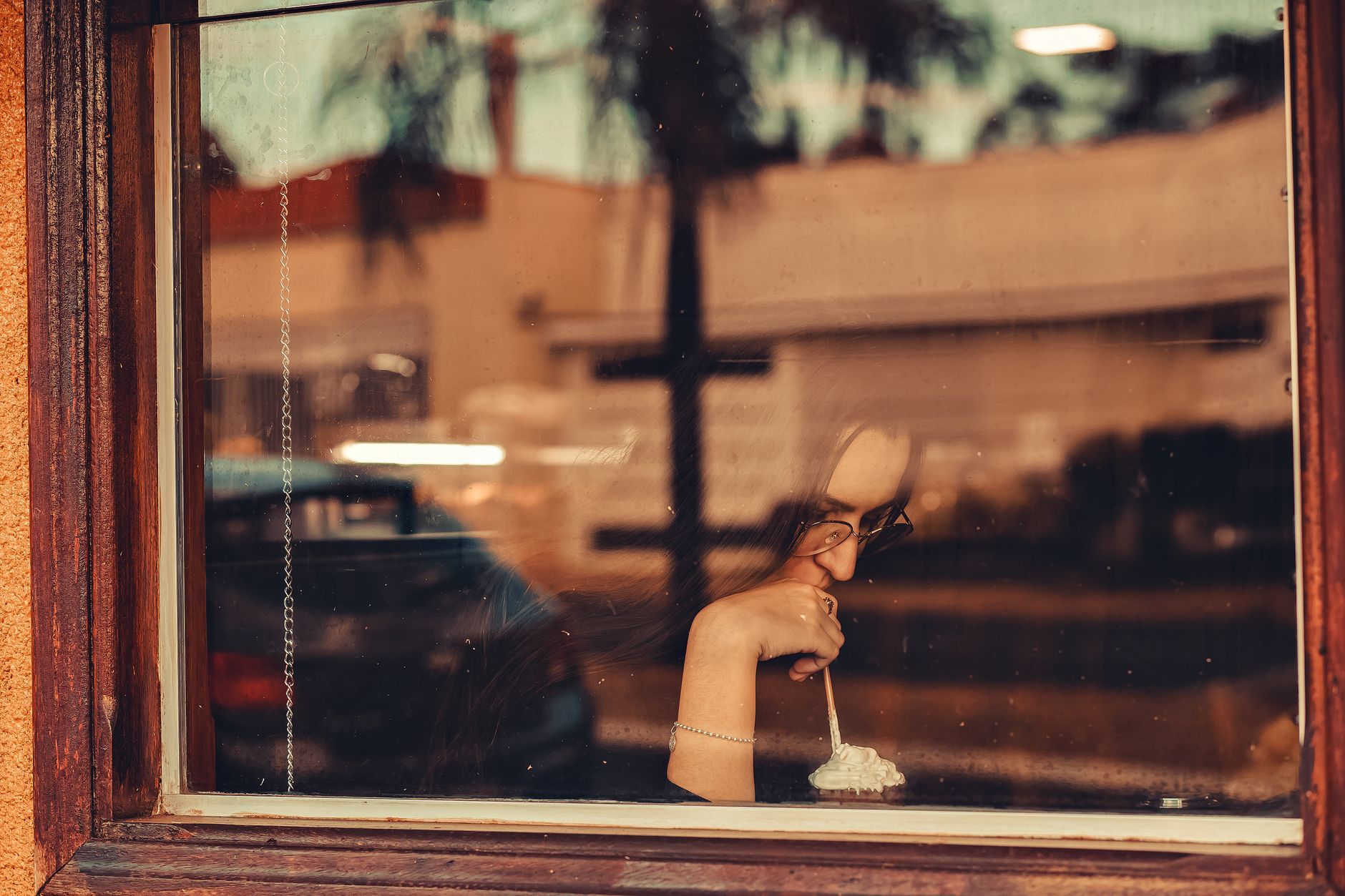 A woman sits by a café window, half-turned toward her drink, thoughtful and a little reserved.