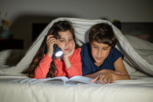 A Girl and a Boy Under a Blanket Reading a Book