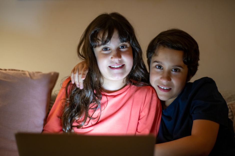 A brother and sister sit closely together using a laptop indoors, smiling warmly.