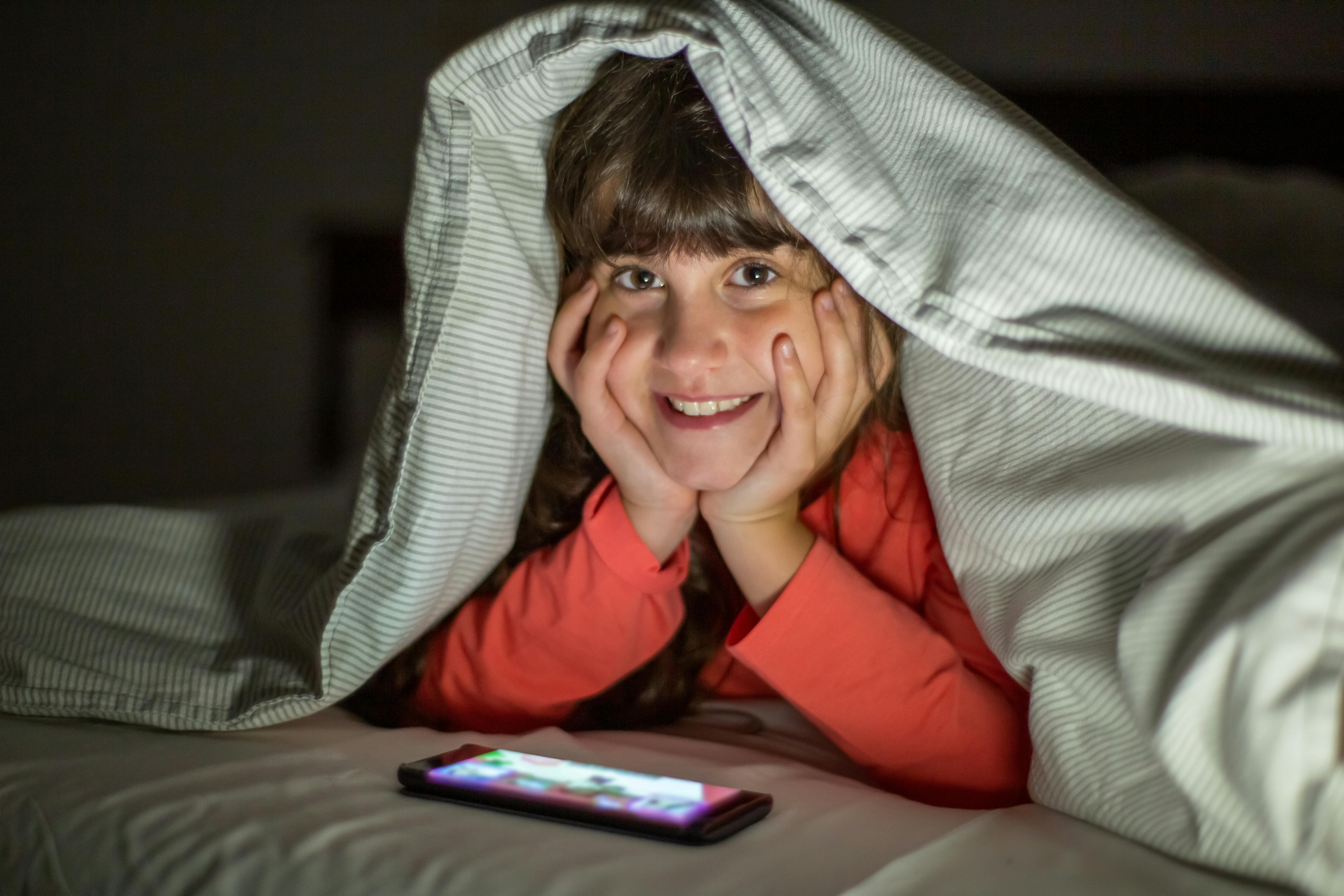 A young girl smiling under a blanket, using a smartphone at night indoors.