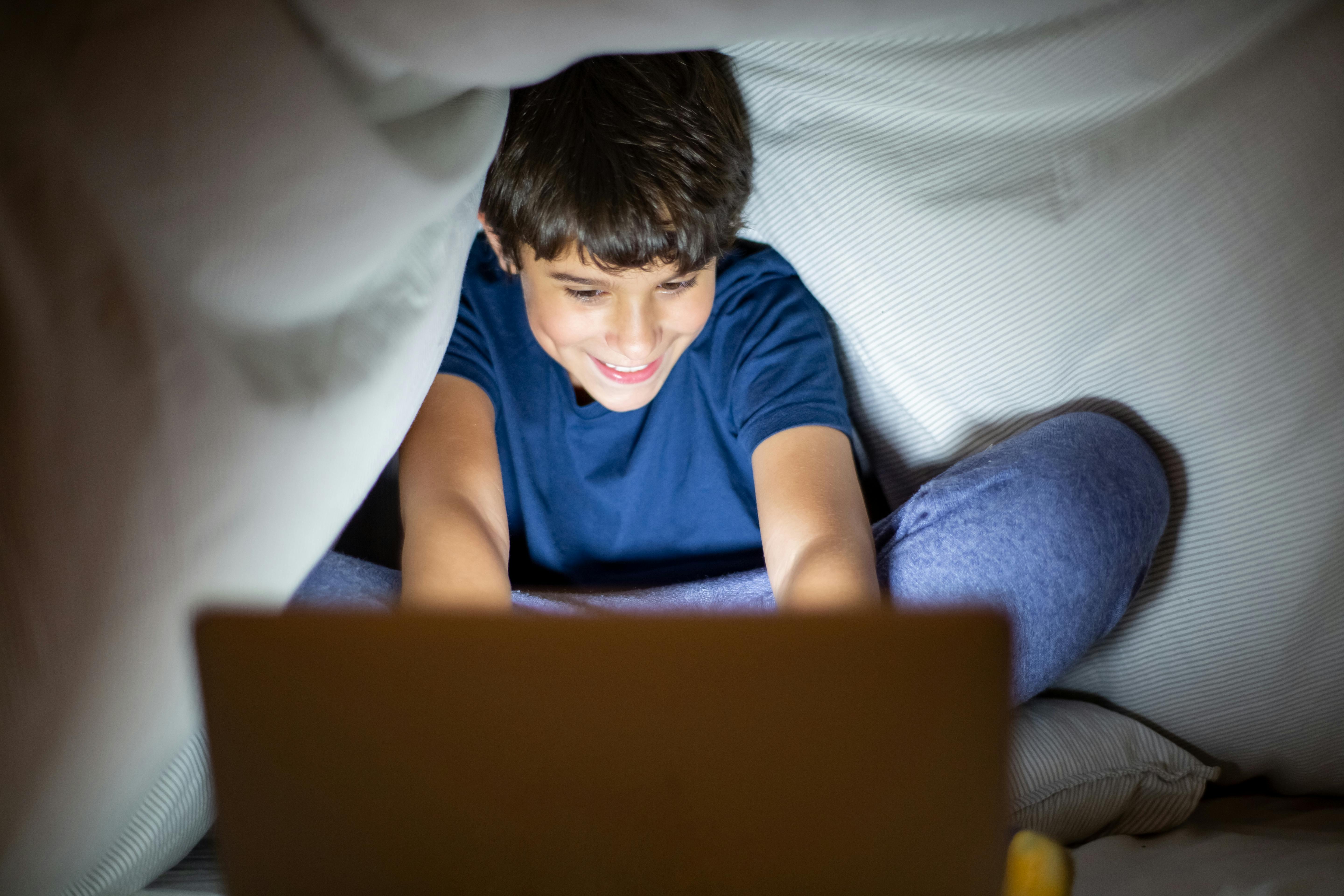 Smiling boy sitting under blanket using laptop, indoor fun at night.