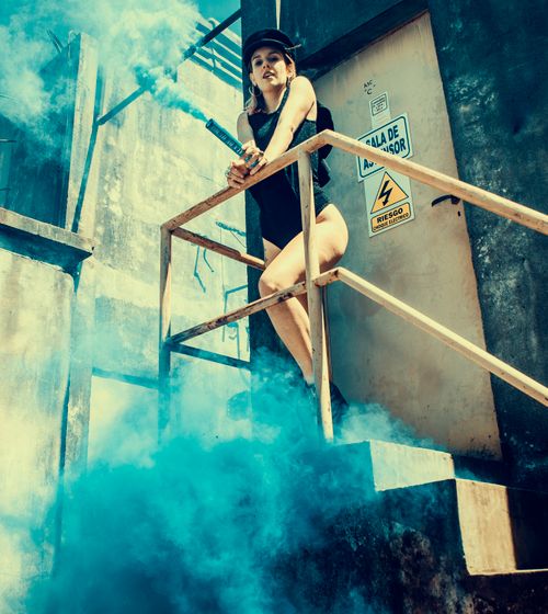 Free Fashionable woman with smoke bomb on industrial stairs in blue haze outdoors. Stock Photo