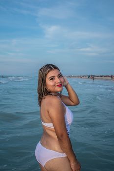 A woman in swimwear poses confidently at the beach with waves and clear sky.