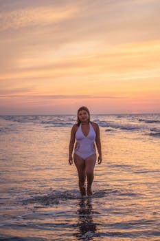 A woman in a swimsuit walking through calm sea waters under a vibrant sunset sky.