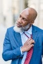 Frustrated bald African American male employee in formal attire loosening tie while standing on street against blurred background after workday