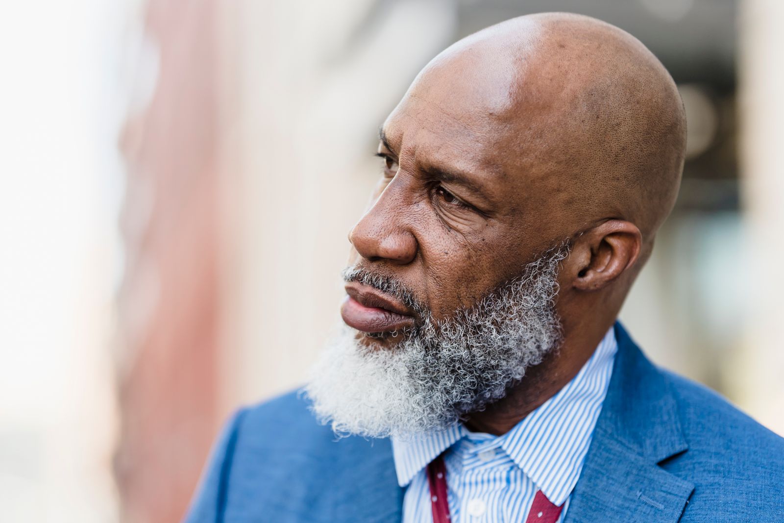 A bald older Black man with a gray-white beard, in a blue blazer and striped shirt, gazes contemplatively into middle distance against a softly blurred urban background.