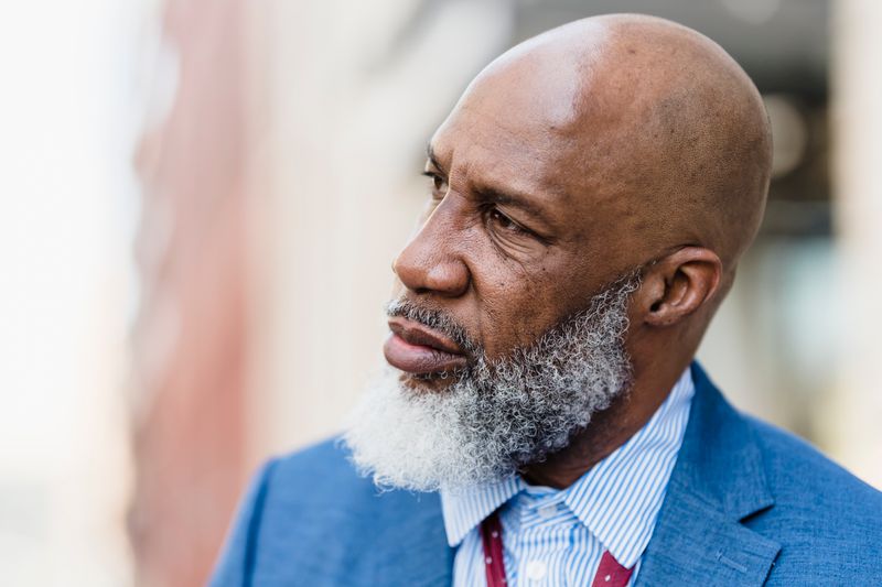 A bald older Black man with a gray-white beard, in a blue blazer and striped shirt, gazes contemplatively into middle distance against a softly blurred urban background.
