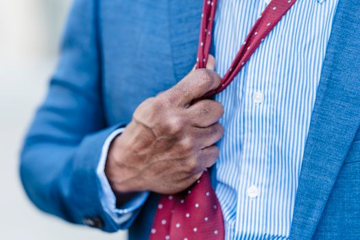 Crop anonymous tired male entrepreneur in formal suit adjusting red tie standing on street in city against blurred background after workday